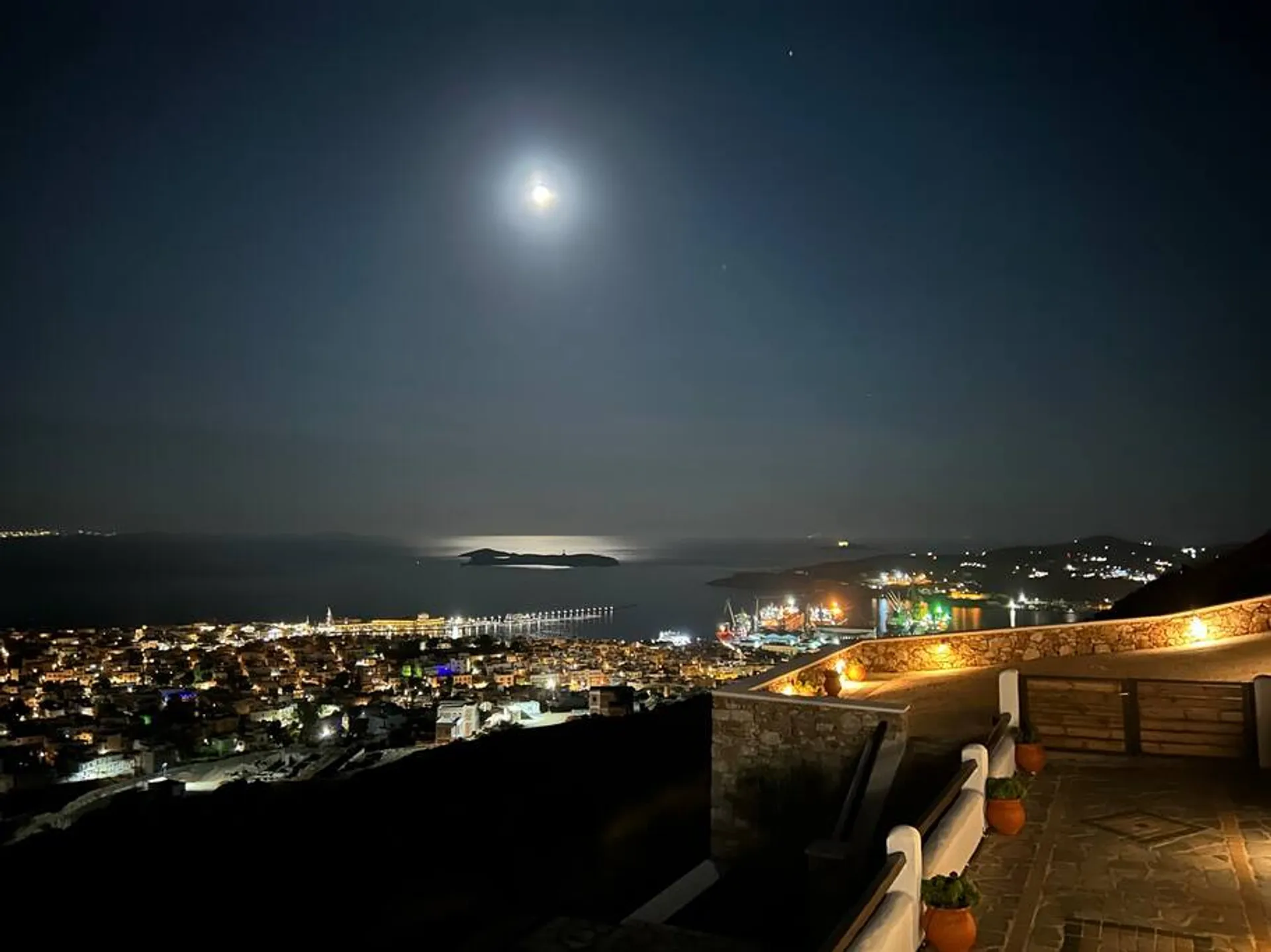 A spectacular night view from the villa's terrace, showing the full moon reflecting on the Aegean Sea and the glittering cityscape of Ermoupolis below.